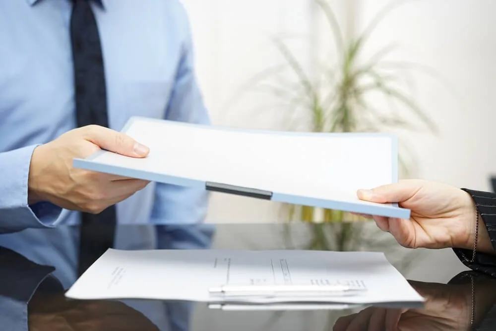 One person handing a stack of documents across a desk to another.